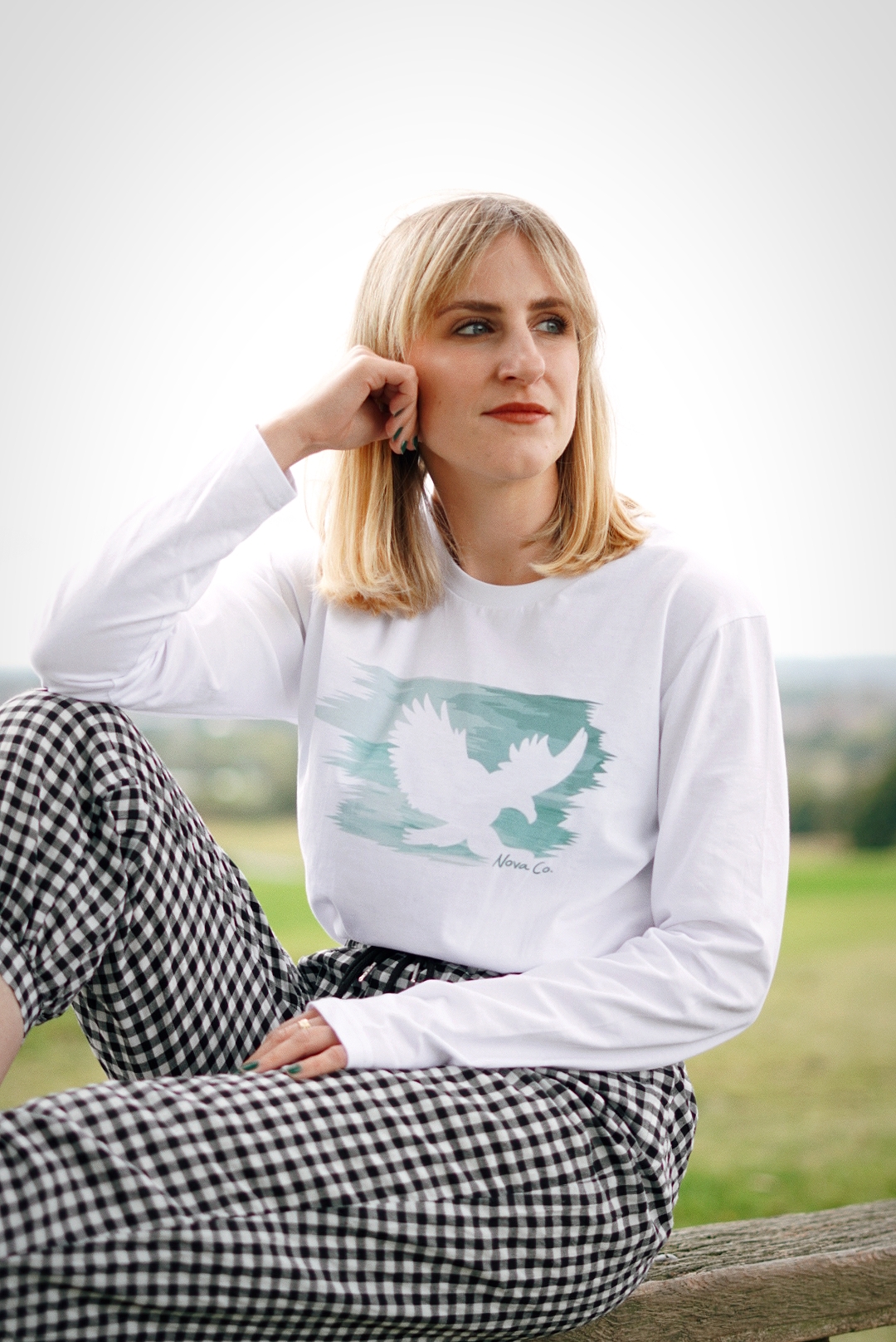 Amy is sat on a bench with her legs up wearing a white long sleeved t-shirt with a graphic of an eagle and gingham trousers